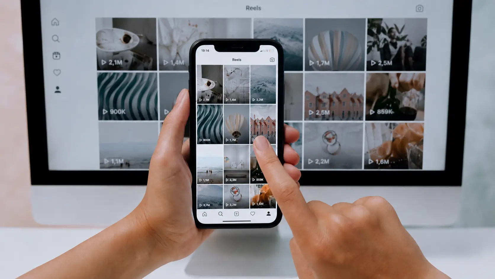 Αλγόριθμος Instagram: Χέρια κρατούν ένα smartphone που εμφανίζει τη σελίδα Reels του Instagram, ενώ στο φόντο μια οθόνη υπολογιστή προβάλλει επίσης ένα grid από Reels.