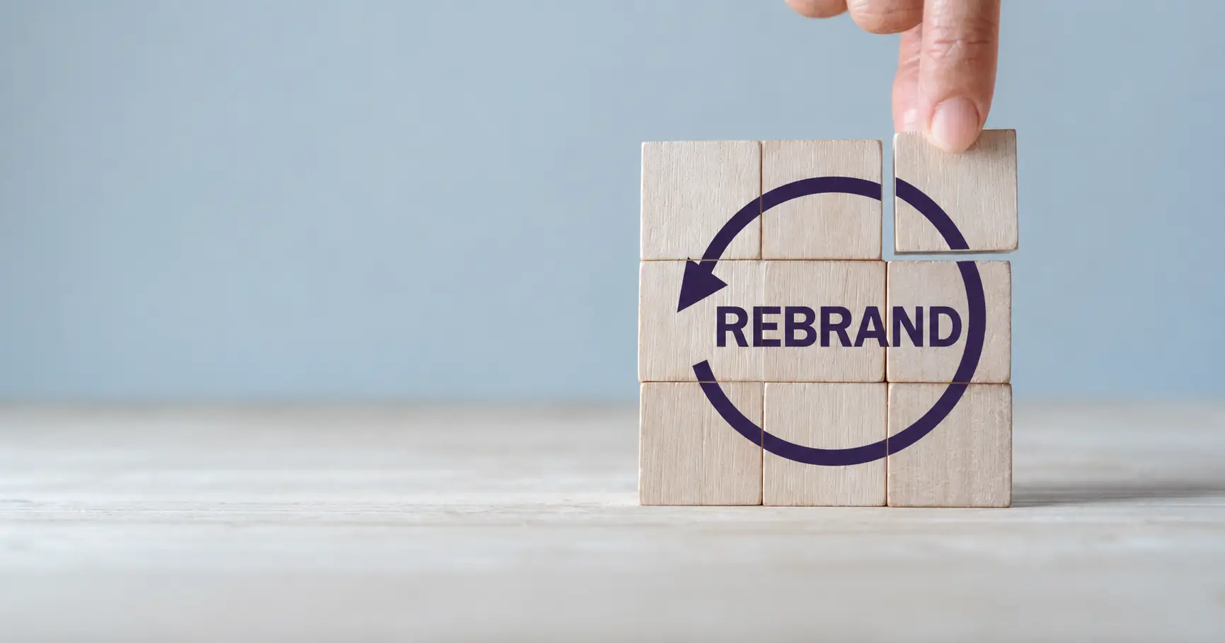 Τι είναι το rebranding: Ξύλινα κύβοι σχηματίζουν τη λέξη «REBRAND» μέσα σε κυκλικό βέλος, ενώ ένα χέρι τοποθετεί ένα κομμάτι του παζλ.